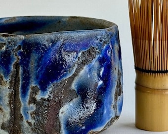 May include: A blue and brown ceramic tea bowl with a textured surface. The bowl is next to a bamboo whisk.