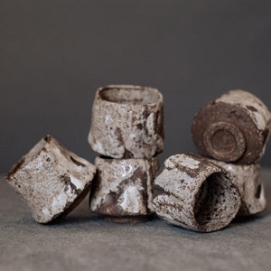 Puede incluir: Un grupo de cinco pequeñas tazas de cerámica con un esmalte blanco moteado y detalles marrones. Las tazas están apiladas y dispuestas en un patrón informal y asimétrico.