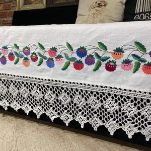 May include: White linen tablecloth with a crocheted lace border. The tablecloth features a colorful embroidered floral design with strawberries and leaves.