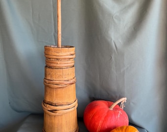 May include: A tall, cylindrical wooden butter churn featuring vertical staves held together by three horizontal wooden bands, with a long, slender wooden plunger extending from the top, positioned next to one large red pumpkin and one smaller orange pumpkin.