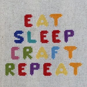 Op de afbeelding: Kruissteekkunstwerk op een neutrale linnen achtergrond. De tekst "EAT SLEEP CRAFT REPEAT" wordt weergegeven in kleurrijke kruissteekletters. De letters zijn in verschillende kleuren, waaronder rood, geel, blauw, groen en paars.