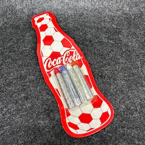 Op de afbeelding: Een rood-witte Coca-Cola-vormige verpakking met een voetbalpatroon, met vier zilverkleurige kleurpotloden met rode, blauwe en oranje punten. Het Coca-Cola-logo is zichtbaar op de verpakking.