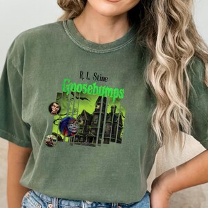 Puede incluir: Una camiseta de manga corta en color verde oliva desteñido con un estampado gráfico de una casa encantada y el texto R.L. Stine Goosebumps en verde brillante. El diseño incluye pequeños retratos circulares y un ambiente oscuro.