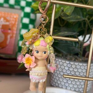 May include: A keychain featuring a small doll with a tropical theme. The doll has a yellow and pink hair, a pink flower, and a skirt with ruffles. The keychain has a gold-colored clasp and decorative beads.