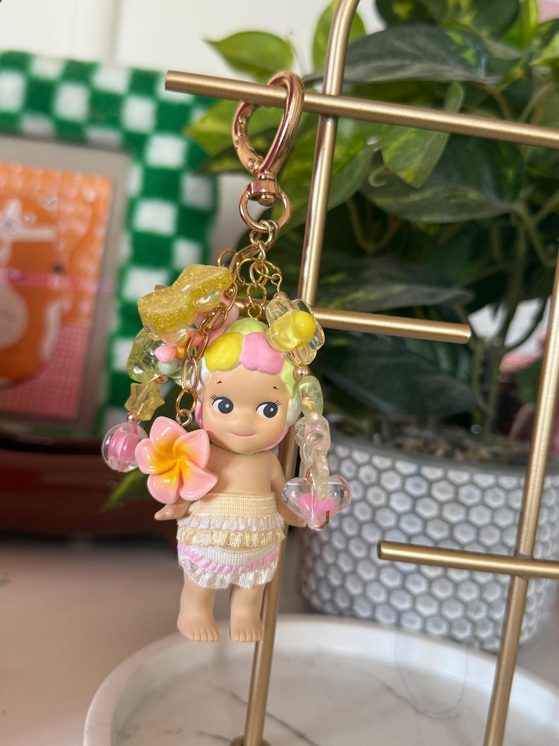 May include: A keychain featuring a small doll with a tropical theme. The doll has a yellow and pink hair, a pink flower, and a skirt with ruffles. The keychain has a gold-colored clasp and decorative beads.