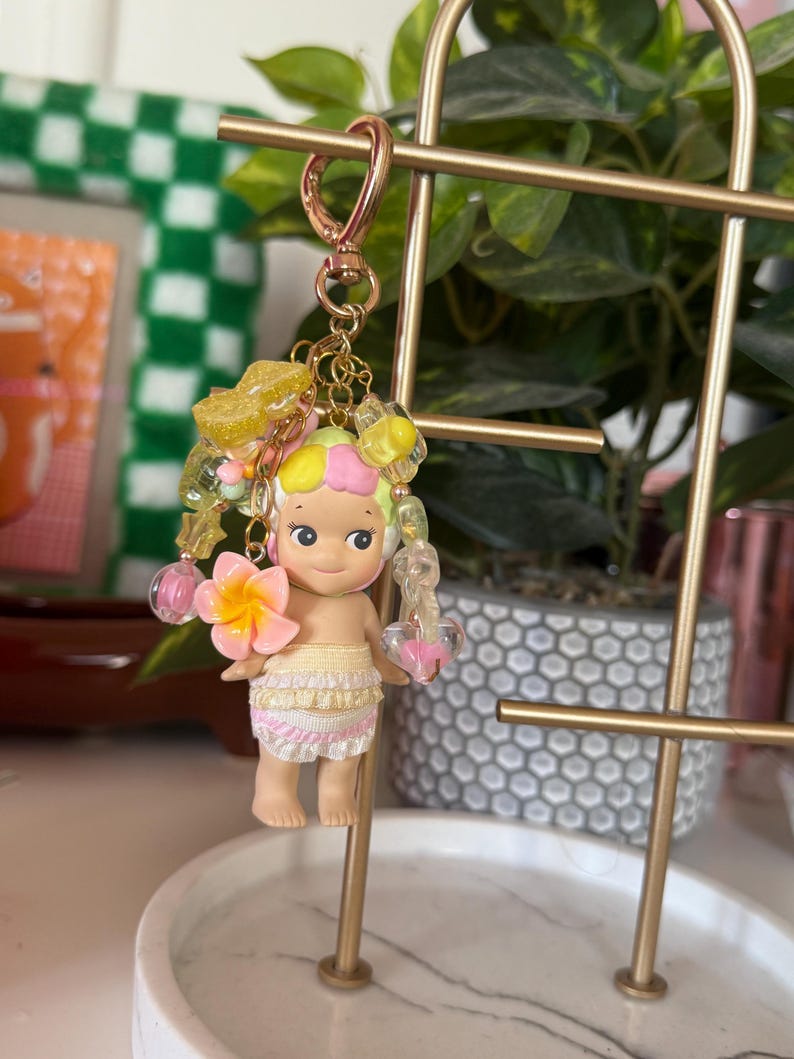 May include: A keychain featuring a small doll with a flower in its hand. The doll has a yellow and pink hair, and a skirt with pink and white ruffles. The keychain has a gold-colored clasp and decorative beads.