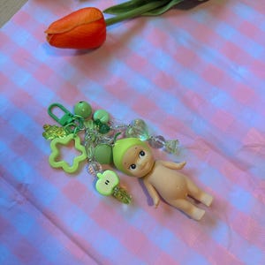Puede incluir: Un llavero con una peque&ntilde;a mu&ntilde;eca y abalorios verdes, incluyendo una estrella, una manzana y cuentas. La mu&ntilde;eca est&aacute; desnuda con un gorro verde. El fondo es un patr&oacute;n de cuadros rosas y blancos, con tulipanes naranjas y amarillos.