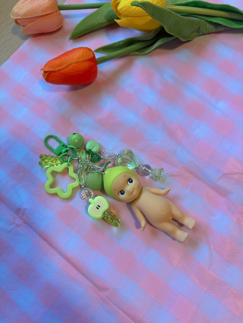 Puede incluir: Un llavero con una peque&ntilde;a mu&ntilde;eca y abalorios verdes, incluyendo una estrella, una manzana y cuentas. La mu&ntilde;eca est&aacute; desnuda con un gorro verde. El fondo es un patr&oacute;n de cuadros rosas y blancos, con tulipanes naranjas y amarillos.