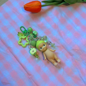 Puede incluir: Un llavero con una peque&ntilde;a mu&ntilde;eca con gorro verde, rodeada de abalorios verdes, incluyendo una estrella, cuentas y una manzana. La mu&ntilde;eca est&aacute; desnuda con una expresi&oacute;n serena. El fondo es un patr&oacute;n de cuadros rosas y blancos con tulipanes naranjas y amarillos.