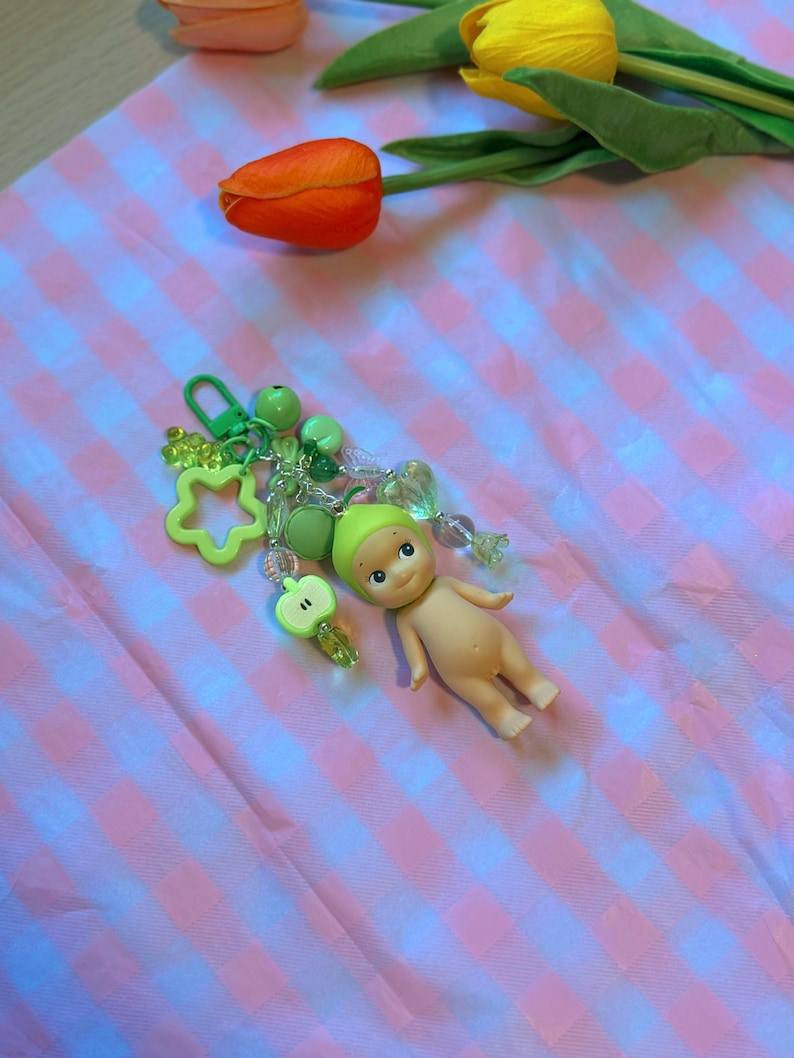 Puede incluir: Un llavero con una peque&ntilde;a mu&ntilde;eca con gorro verde, rodeada de abalorios verdes, incluyendo una estrella, cuentas y una manzana. La mu&ntilde;eca est&aacute; desnuda con una expresi&oacute;n serena. El fondo es un patr&oacute;n de cuadros rosas y blancos con tulipanes naranjas y amarillos.