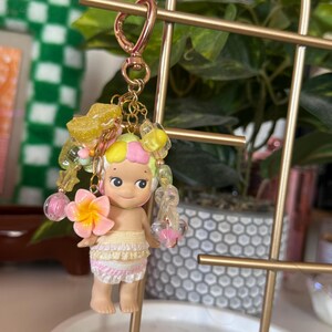 May include: A keychain featuring a small doll with a tropical theme. The doll has a yellow and pink hair, a flower, and a skirt. The keychain includes a gold-colored clasp and decorative beads in yellow, pink, and clear colors.