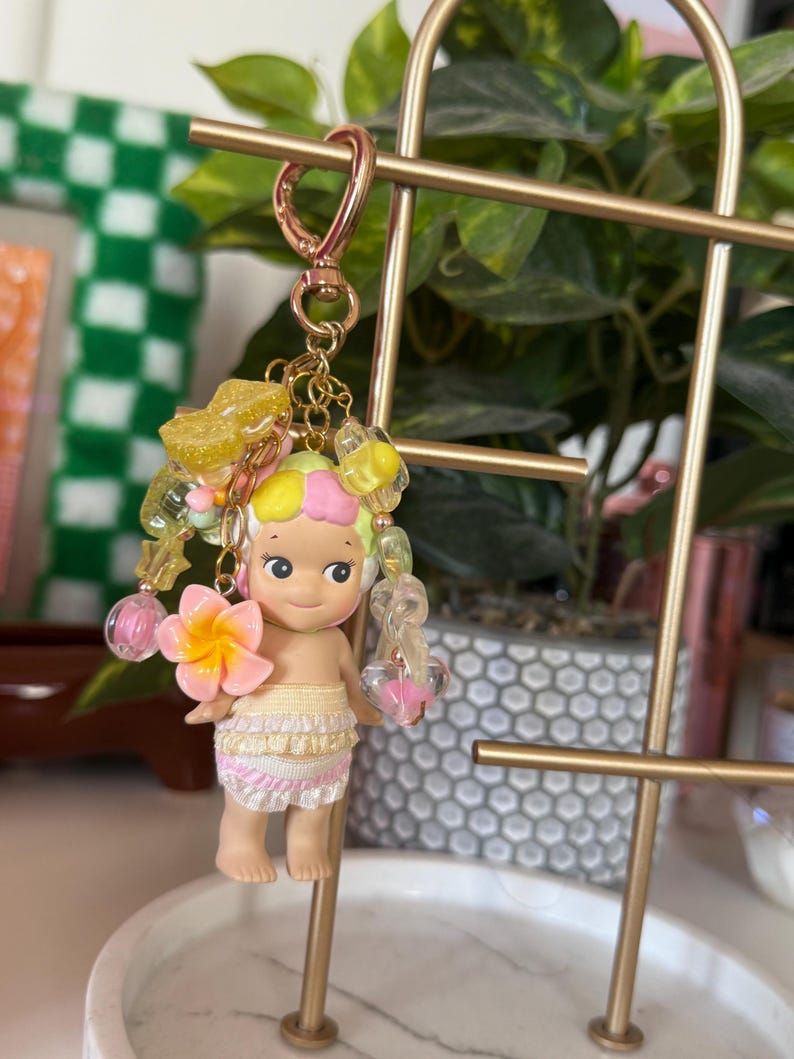 May include: A keychain featuring a small doll with a tropical theme. The doll has a yellow and pink hair, a flower, and a skirt. The keychain includes a gold-colored clasp and decorative beads in yellow, pink, and clear colors.
