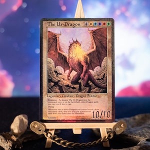 Könnte beinhalten: Sammelkarte mit einer Drachenillustration und dem Text "The Ur-Dragon". Die Karte wird auf einer kleinen Holzstaffelei präsentiert, umgeben von Steinen, vor einem farbenfrohen Hintergrund.