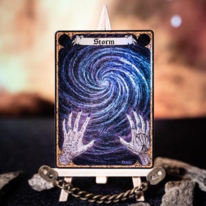 Può includere: Una carta d'arte fantasy intitolata "Storm" esposta su un cavalletto in miniatura. La carta presenta un disegno a spirale blu e bianco con mani tese. La parola "Storm" è nella parte superiore della carta.
