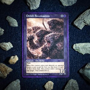 May include: A collectible trading card titled "Orcish Bowmasters" with a purple border. The card features a detailed illustration of a monstrous creature. The card is surrounded by various gray stones on a dark surface.