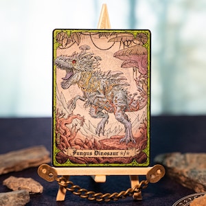 Puede incluir: Una tarjeta de arte de fantasía que presenta un dinosaurio con crecimientos en forma de hongo, etiquetada como "Fungus Dinosaur". La tarjeta se muestra en un caballete de madera en miniatura con una cadena dorada. La obra de arte tiene un borde verde y un estilo de ilustración detallado.
