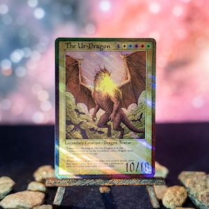 Op de afbeelding: Een holografische Magic: The Gathering ruilkaart getiteld The Ur-Dragon, met een grote bruine draak met gespreide vleugels, een goudkleurige gloeiende kop en kleinere vliegende draken op de achtergrond, met 10/10 kracht en weerstand.