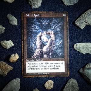 Op de afbeelding: Een Mox Opal-kaart, een legendarisch artefact, met een illustratie van een gloeiende bol die door handen wordt vastgehouden. De kaart is omgeven door rotsen op een donkerblauwe ondergrond. De tekst op de kaart luidt "Metalcraft - Voeg één mana van elke kleur toe."
