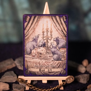 Puede incluir: Una carta de arte de fantasía que representa ratas reunidas alrededor de una mesa con una calavera y velas. La carta tiene un borde púrpura y dice "this token can't block" y "Rat 1/1". Se muestra en un caballete de madera en miniatura.