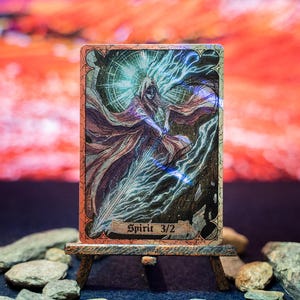 Pode incluir: Uma carta colecionável retangular com uma figura encapuzada em mantos fluidos empunhando uma espada brilhante, com um efeito de luz circular brilhante atrás. A carta exibe o texto Spirit 3/2 na parte inferior e está em um pequeno cavalete.