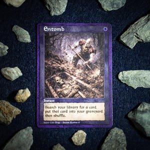 May include: A Magic: The Gathering card titled "Entomb" with a dark purple border. The card features an illustration of a figure digging a grave. The text reads "Search your library for a card, put that card into your graveyard, then shuffle."