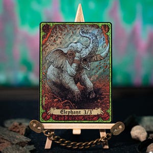 Puede incluir: Una ilustración detallada de un elefante en una tarjeta, exhibida en un caballete de madera en miniatura. La tarjeta presenta el texto "Elephant 3/3" y un borde verde con detalles florales. El elefante es gris con grandes colmillos.