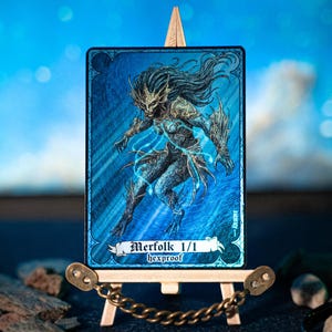May include: A fantasy art card featuring a blue-skinned mermaid-like creature with long dark hair and intricate details. The card is displayed on a small wooden easel with the text "Merfolk 1/1 hexproof". The background is a blurred blue.