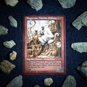 May include: A Magic: The Gathering card, "Ragavan, Nimble Pilferer," with a red border and a detailed illustration of a white monkey. The card is surrounded by small gray rocks on a dark blue surface. Text on the card describes its abilities.