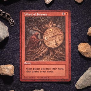 Puede incluir: Una carta roja "Wheel of Fortune" con una ilustración detallada de una figura con túnica girando una rueda de madera. El texto de la carta dice: "Cada jugador descarta su mano, luego roba siete cartas." La carta está rodeada de piedras.