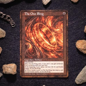Op de afbeelding: Een ruilkaart met "The One Ring", een gouden ring met gegraveerde tekst, tegen een vurige achtergrond. De kaart heeft een bruine rand. Kleine stenen omringen de kaart op een donkere ondergrond. De kaart luidt "Legendary Artifact".