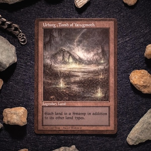Könnte beinhalten: Eine Magic: The Gathering-Karte mit dem Titel "Urborg, Tomb of Yawgmoth", die eine düstere, atmosphärische Illustration einer sumpfigen Landschaft unter einem stürmischen Himmel zeigt. Die Karte ist von kleinen Steinen auf einer dunklen Oberfläche umgeben. Der Text auf der Karte lautet "Legendary Land".
