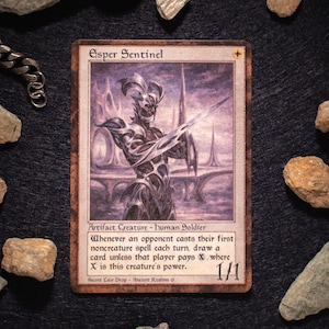May include: A Magic: The Gathering card featuring the "Esper Sentinel." The card depicts a humanoid figure with a sword, set against a purple and white background. The card's text includes "Artifact Creature - Human Soldier" and other game instructions. The card is surrounded by rocks.