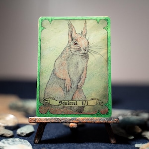 Pode incluir: Um cartão colecionável retangular com uma ilustração detalhada a tinta e aguarela de um esquilo em pé. O cartão tem uma borda verde vibrante e uma faixa na parte inferior com o texto preto Squirrel 1/1 impresso nele.