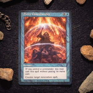 Può includere: Una carta di Magic: The Gathering intitolata "Fierce Guardianship" con bordo blu. L'artwork raffigura un mago con un bastone, circondato da uno sfondo infuocato, e figure sottostanti. Il testo della carta include "Instant" e i dettagli dell'incantesimo.