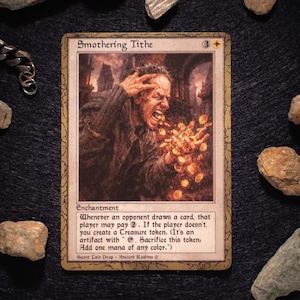Può includere: Una carta di Magic: The Gathering intitolata "Smothering Tithe" con un'illustrazione di un uomo con monete. La carta ha un bordo dorato e testo di gioco. La carta è circondata da rocce.