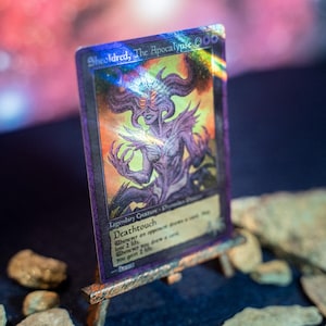 Op de afbeelding: Een holografische ruilkaart getiteld Sheoldred, The Apocalypse, met een paarsgetinte illustratie van een gehoornd wezen met gloeiende ogen. De kaart toont de tekst Deathtouch en 4/6 en staat op een kleine houten ezelstandaard.