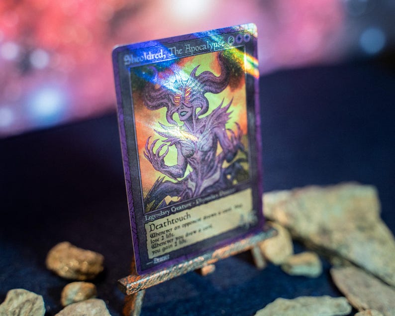 Op de afbeelding: Een holografische ruilkaart getiteld Sheoldred, The Apocalypse, met een paarsgetinte illustratie van een gehoornd wezen met gloeiende ogen. De kaart toont de tekst Deathtouch en 4/6 en staat op een kleine houten ezelstandaard.