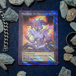 Op de afbeelding: Een holografische Sheoldred, The Apocalypse ruilkaart met een wezen met meerdere hoorns en gloeiende ogen, omringd door verspreide grijze stenen, een zilveren ketting en een donkere metalen ring op een zwarte ondergrond met speltekst.