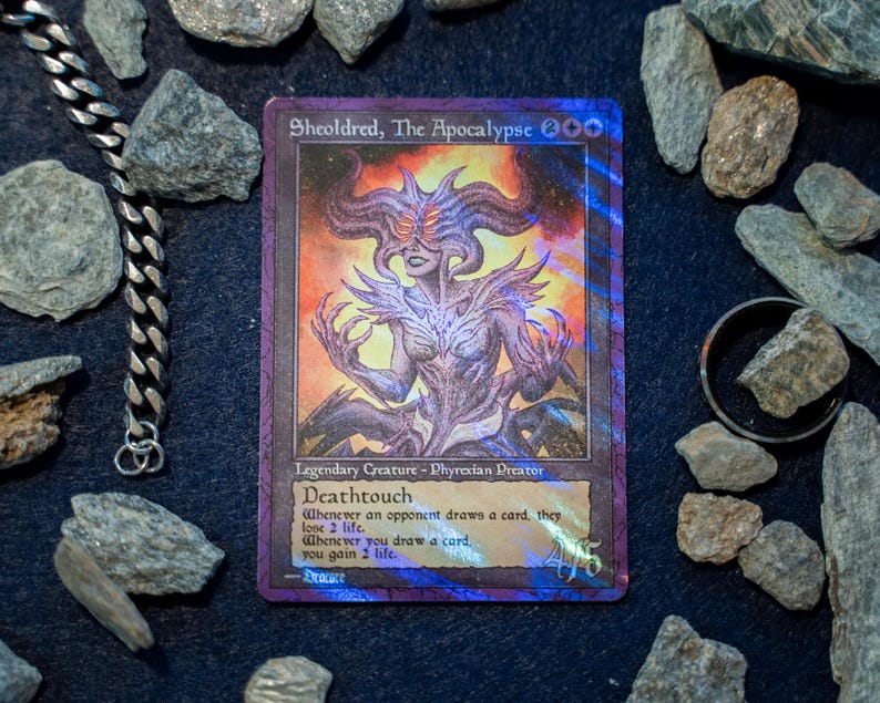 Op de afbeelding: Een holografische Sheoldred, The Apocalypse ruilkaart met een wezen met meerdere hoorns en gloeiende ogen, omringd door verspreide grijze stenen, een zilveren ketting en een donkere metalen ring op een zwarte ondergrond met speltekst.