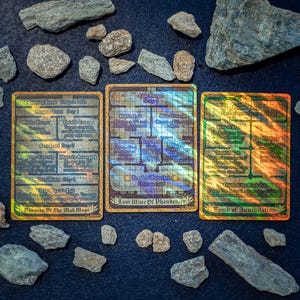 Peut inclure: Trois cartes de jeu rectangulaires holographiques avec des grilles et du texte. La carte de gauche est intitulée Dungeon Of The Mad Mage, celle du centre Lost Mine Of Phandelver, et celle de droite Tomb Of Annihilation, entourées de rochers gris.