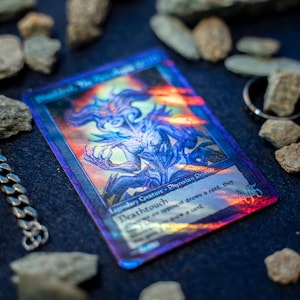 Op de afbeelding: Een holografische ruilkaart met een paarse en blauwe illustratie van een legendarisch wezen, genaamd Sheoldred, the Apocalypse, met de tekst Deathtouch en Phyrexian Praetor, omringd door verspreide grijze stenen en een metalen ketting.
