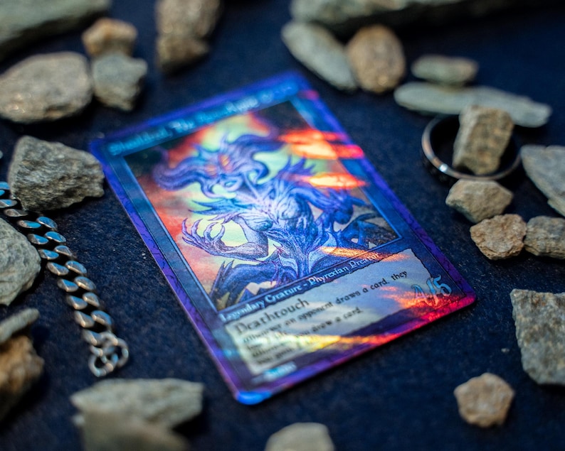 Op de afbeelding: Een holografische ruilkaart met een paarse en blauwe illustratie van een legendarisch wezen, genaamd Sheoldred, the Apocalypse, met de tekst Deathtouch en Phyrexian Praetor, omringd door verspreide grijze stenen en een metalen ketting.