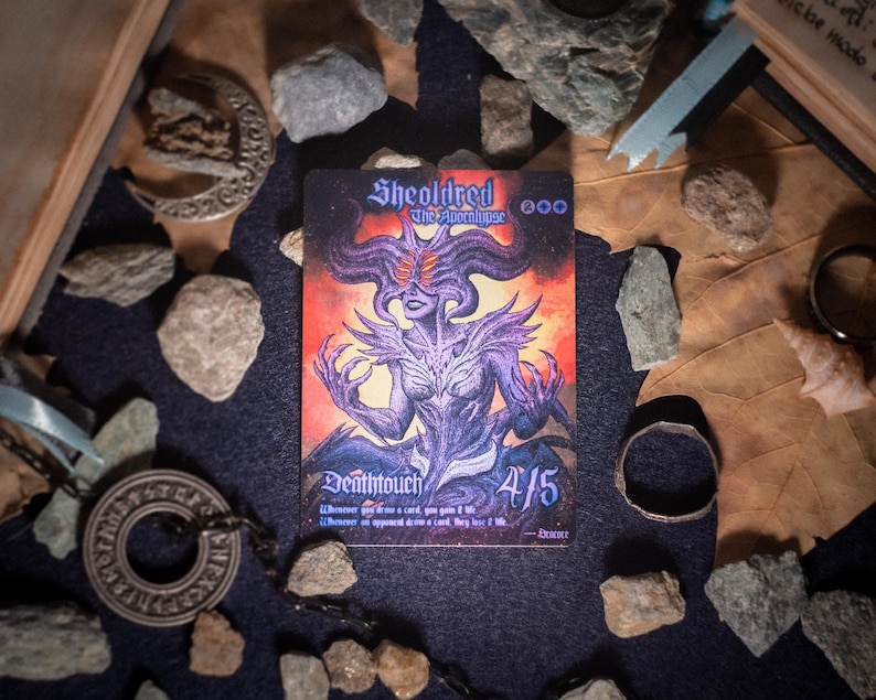 Op de afbeelding: Een rechthoekige ruilkaart met een paarse, gehoornde figuur met gloeiende ogen tegen een vurige oranje en zwarte achtergrond. De tekst luidt Sheoldred The Apocalypse, Deathtouch, 4/5, en bevat spelregels onderaan de kaart.
