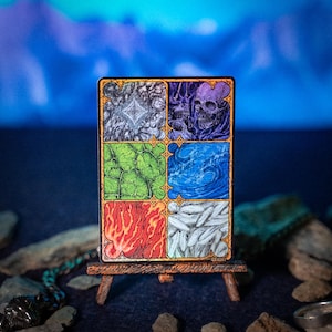 Può includere: Una carta rettangolare illustrata con sei pannelli raffiguranti elementi diversi: un teschio, acqua, fuoco, ghiaccio e altri disegni astratti. La carta è esposta su un piccolo cavalletto di legno su uno sfondo blu sfocato.