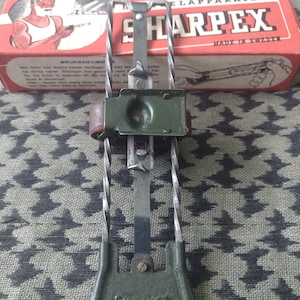 Puede incluir: Afilador de cuchillas de afeitar SHARPEX vintage en verde y plateado, con una caja roja en el fondo. El afilador tiene un mango y un marco de metal. La caja presenta el texto "SHARPEX MADE IN SWEDEN" y un dibujo de un hombre afeitándose.
