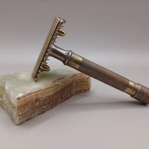 May include: An antique safety razor with a textured handle and a gold-colored head. The razor is resting on a light green and brown stone. The razor's design suggests it is a vintage grooming tool.