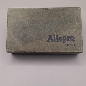 Può includere: Una scatola rettangolare in cartone grigio usurato con una superficie ruvida. La parte superiore presenta la scritta blu scuro Allegro e le lettere MOD. L stampate nell'angolo in basso a destra. La scatola ha un aspetto vissuto con graffi.