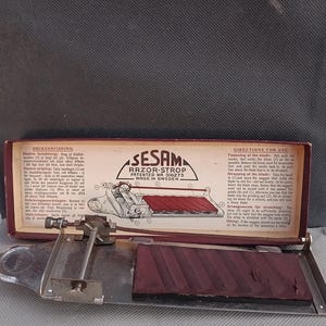 Puede incluir: Un afilador de navajas vintage Sesam con una tira de cuero roja y un marco de metal. La caja está abierta y las instrucciones de uso son visibles. La caja está etiquetada "Sesam Razor-Strop Patentado Nr 516273 Hecho en Suecia."
