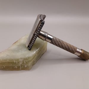 May include: A vintage safety razor with a silver head and a textured, silver and gold handle rests on a light green, layered stone. The razor is a classic grooming tool.
