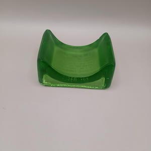 May include: A green glass ashtray with a curved design. The ashtray has a rectangular base and a curved top, with a smooth, glossy finish. The glass is a vibrant shade of green, and the ashtray appears to be made of thick glass.